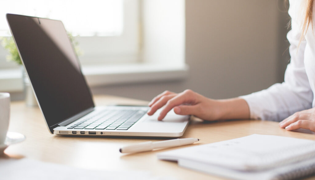 Zdjęcie laptopa i dokumentów leżących na biurku, widoczna część kobiecej sylwetki pracującej na laptopie